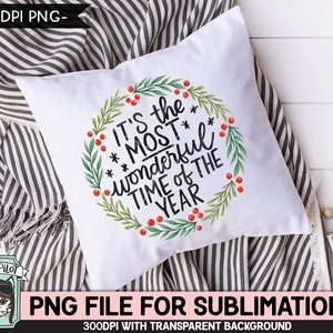 May include: White square pillow with the text "It's the MOST wonderful TIME OF THE YEAR" in black script, surrounded by a green wreath with red berries. The pillow is on a striped blanket.