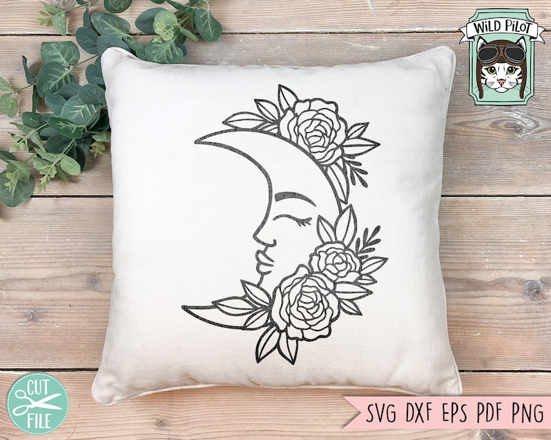 May include: A white pillow with a black line drawing of a crescent moon with a face and floral details. The moon is surrounded by flowers and leaves.