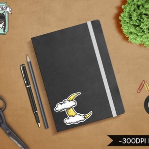 May include: A black notebook with a white elastic band closure. A yellow crescent moon sticker with white clouds is on the front cover. The notebook is on a brown surface with a pair of scissors, two pens, and paper clips.
