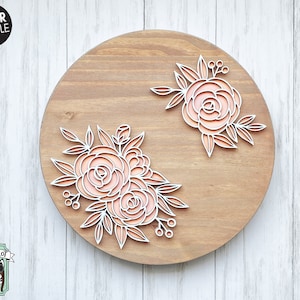 Puede incluir: Una placa redonda de madera con dos dise&ntilde;os florales cortados con l&aacute;ser. Las flores son de color melocot&oacute;n y el dise&ntilde;o es blanco. La placa est&aacute; sobre un fondo de madera blanca.