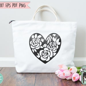 May include: A white tote bag with a black floral heart design. The heart is filled with stylized flowers and leaves. The bag is sitting on a wooden surface with a bouquet of pink tulips in the foreground.