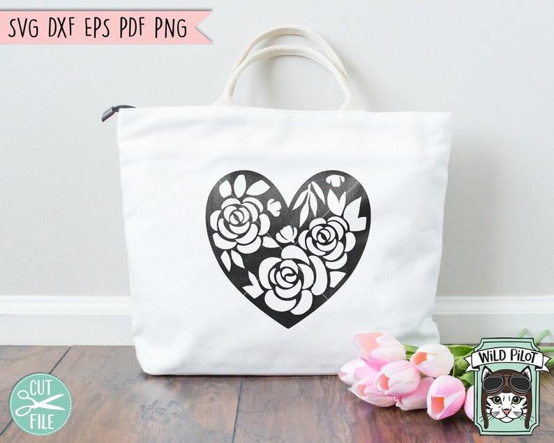 May include: A white tote bag with a black floral heart design. The heart is filled with stylized flowers and leaves. The bag is sitting on a wooden surface with a bouquet of pink tulips in the foreground.