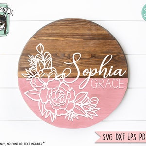 May include: A round wooden sign with a pink and brown background. The top half of the sign is brown and the bottom half is pink. The sign has a white floral design and the text "Sophia Grace" in white cursive lettering.