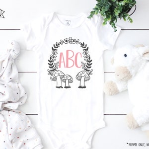 Op de afbeelding: Een witte romper met een zwart-witte kransontwerp met paddenstoelen en de letters ABC in roze. De romper ligt op een witte houten achtergrond.