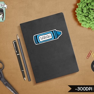 Peut inclure: Un carnet noir avec un autocollant de crayon bleu qui dit "SARAH" dessus. Le carnet est sur une surface brune avec une paire de ciseaux, deux stylos et quelques trombones.