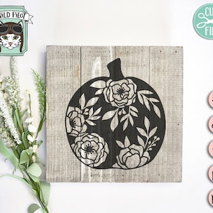Op de afbeelding: Een zwart silhouet van een pompoen met bloemdetails op een houten achtergrond. De pompoen is omringd door witte bloemen en bladeren.