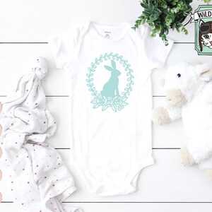 May include: A white baby onesie with a teal bunny silhouette inside a wreath of flowers. The onesie is on a white wooden surface with a white stuffed animal and a pink and white polka dot blanket.