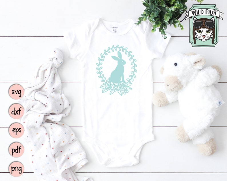 May include: A white baby onesie with a teal bunny silhouette inside a wreath of flowers. The onesie is on a white wooden surface with a white stuffed animal and a pink and white polka dot blanket.