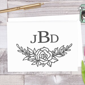 May include: A black and white monogram design with the letters "JBD" surrounded by a floral wreath. The wreath is made up of leaves and a single rose.