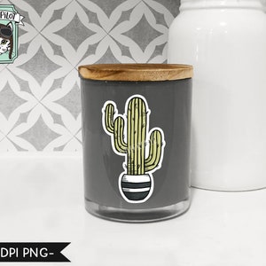May include: A gray candle jar with a wooden lid and a cactus sticker on the front. The sticker features a green cactus in a black and white striped pot.