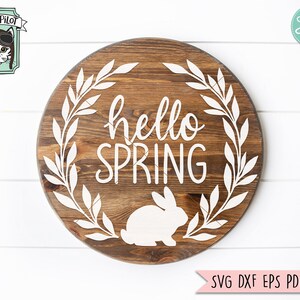 Op de afbeelding: Een rond houten bord met een witte krans en de tekst "hello spring" in witte letters. Een wit silhouet van een konijn bevindt zich in het midden van het bord.