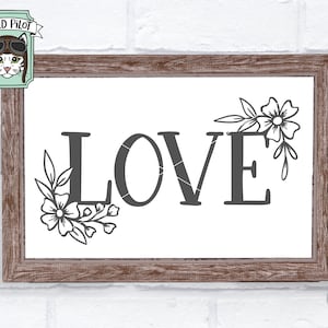 May include: A white sign with a brown wooden frame, featuring the word "LOVE" in a bold, black font. The word is adorned with delicate floral designs on either side, adding a touch of elegance. The sign is perfect for a romantic setting or as a decorative piece for a home.