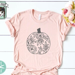 Op de afbeelding: Een roze T-shirt met een zwart bloemig pompoenontwerp. Het ontwerp is zwart omlijnd en bevat een steel en bladeren. Het shirt is een populaire keuze voor herfstmode.