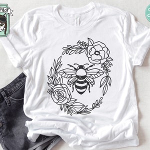 Puede incluir: Una camiseta blanca con un dise&ntilde;o en blanco y negro de una abeja rodeada de flores. El dise&ntilde;o tiene forma circular.