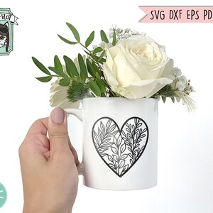 May include: A white ceramic mug with a black outline of a heart filled with leafy branches. The mug is holding a bouquet of white roses and greenery.
