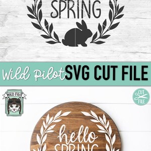 Op de afbeelding: Een digitaal snijbestand met een krans van bladeren met de tekst "hello spring" en een konijnen silhouet. Het ontwerp is weergegeven op een houten achtergrond.