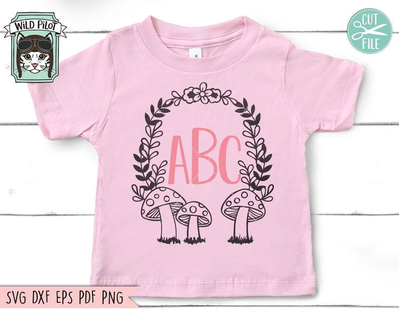 Op de afbeelding: Een roze baby-onesie met een zwart-witte krans met de letters ABC in roze erin. Er zijn drie zwart-witte paddenstoelen onderaan de krans.