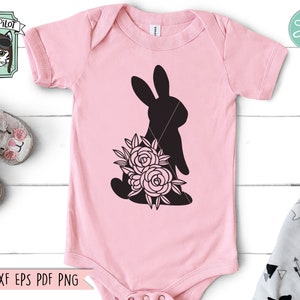 Op de afbeelding: Een lichtroze babyromper met een zwart silhouet van een konijn met bloemen. De romper heeft korte mouwen en een ronde hals. De tekst "SVG DXF EPS PDF PNG" staat onderaan. Een "Wild Pilot"-logo staat linksboven.
