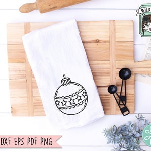 Op de afbeelding: Een witte keukendoek met een zwart-witte omtrek van een kerstbal met sterren. De doek ligt op een houten snijplank met een deegroller en maatschepjes.