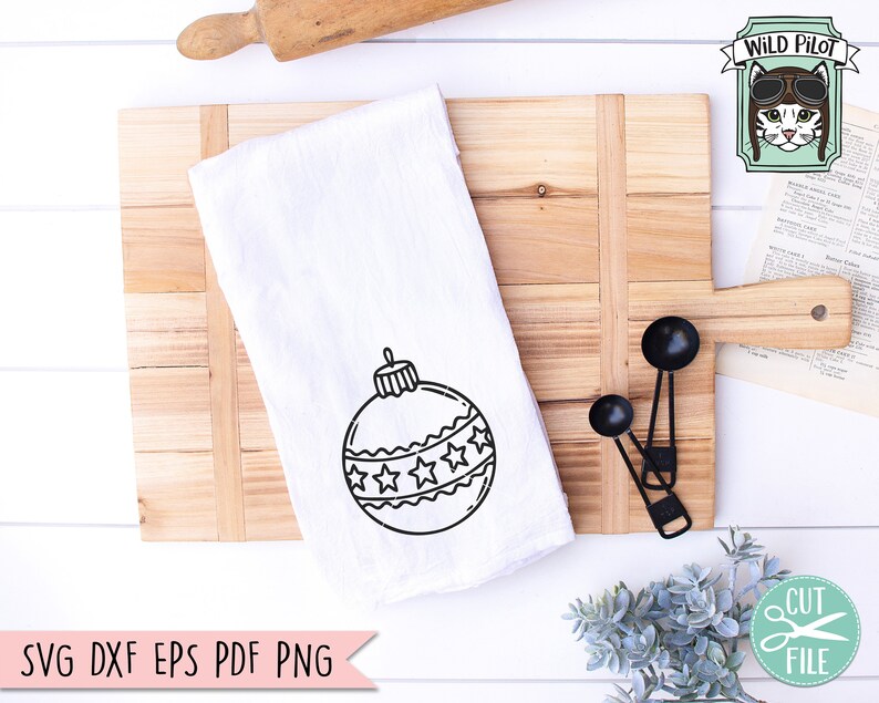 Op de afbeelding: Een witte keukendoek met een zwart-witte omtrek van een kerstbal met sterren. De doek ligt op een houten snijplank met een deegroller en maatschepjes.