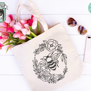 Puede incluir: Una bolsa de tela blanca con una ilustraci&oacute;n en blanco y negro de una abeja rodeada de flores. La abeja est&aacute; en el centro del dise&ntilde;o y tiene un cuerpo rayado. Las flores est&aacute;n dispuestas en un c&iacute;rculo alrededor de la abeja. La bolsa de tela est&aacute; sobre una superficie de madera blanca.