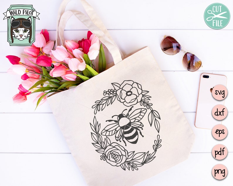 Puede incluir: Una bolsa de tela blanca con una ilustraci&oacute;n en blanco y negro de una abeja rodeada de flores. La abeja est&aacute; en el centro del dise&ntilde;o y tiene un cuerpo rayado. Las flores est&aacute;n dispuestas en un c&iacute;rculo alrededor de la abeja. La bolsa de tela est&aacute; sobre una superficie de madera blanca.