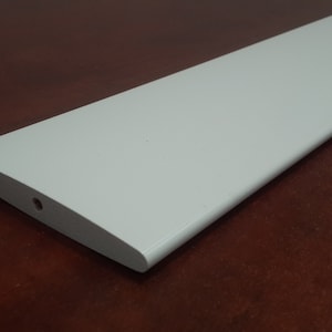 May include: A white, rectangular, flat slat for a window blind. The slat has a rounded edge and a small hole in the center.