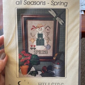May include: A cross-stitch pattern featuring a snowman wearing a green apron and a straw hat. The snowman is holding a watering can and there are flowers and a bird in the background. The pattern is titled "A "Snow" Woman for all Seasons - Spring" and is from Hillside Samplings.