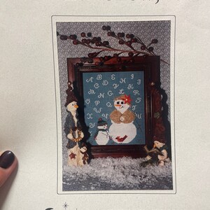 May include: A cross-stitch pattern featuring a framed alphabet sampler with three snowmen. The sampler is on a blue background with white letters. The snowmen are wearing hats and scarves. The text "Let-her Snow, too." is at the top of the pattern. The text "HILLSIDE SAMPLINGS" is at the bottom of the pattern.