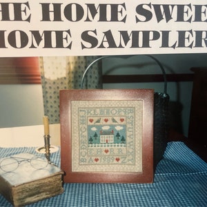 Puede incluir: Un sampler de punto de cruz enmarcado con el texto "The Home Sweet Home Sampler" y el alfabeto en un esquema de color azul y blanco. El sampler presenta una casa con una puerta roja, dos pájaros y nubes. El sampler está sobre un mantel a cuadros azul y blanco.