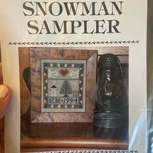 May include: A cross-stitch sampler pattern titled "The Snowman Sampler" by Ramsgate Limited. The pattern features a snowman, a Christmas tree, and two birds with a heart. The pattern is framed in a rustic wooden frame and displayed on a wooden surface with an antique oil lamp.