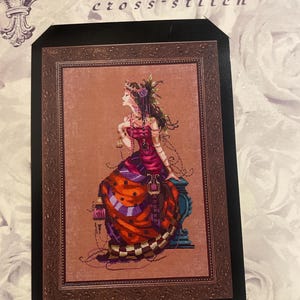 Può includere: Un modello a punto croce incorniciato con una donna in un lungo abito con dettagli arancioni, viola e rossi. L'opera è intitolata "Mirabilia cross-stitch". La donna indossa gioielli e un copricapo floreale.