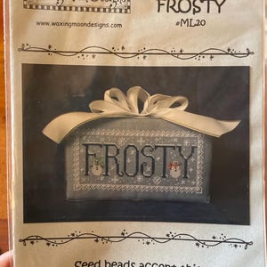 May include: A cross-stitch pattern for a winter-themed design called "Frosty". The pattern features a blue background with white snowflakes and the word "Frosty" in black letters. Two small snowmen are stitched on either side of the word. The pattern is framed by a white ribbon with a bow. The text "Seed beads accent this quick-to-stitch tribute to winter." is printed below the pattern.