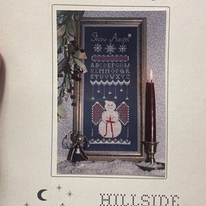 May include: Cross stitch pattern for a winter scene with a snow angel, snowflakes, and the text "Snow Angel". The pattern includes an alphabet and numbers. The pattern is from Hillside Samplings, 1927 Lapham Rd., East Aurora, NY 14052, (716) 655-4091, Designs by Lesley Rudnicki.