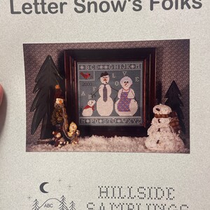 May include: A cross-stitch pattern featuring a family of snowmen. The pattern is titled "Letter Snow's Folks" and is from Hillside Samplings. The pattern includes the alphabet and numbers.