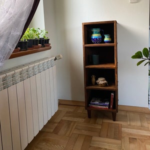 May include: A wooden bookshelf with four shelves. The top shelf has two blue and green ceramic pitchers. The second shelf has a green mug. The third shelf has a small brown bottle and a glass jar with a lid. The bottom shelf has a stack of books. The bookshelf has a dark wood finish and is standing on four legs.