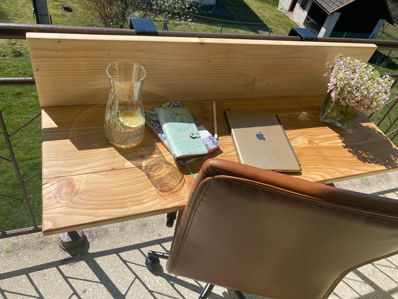 May include: A wooden desk with a brown leather chair, a glass pitcher of water, a tablet, and two notebooks. The desk is attached to a balcony railing.