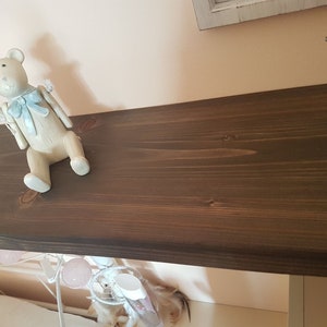 Puede incluir: Una figura decorativa de oso de peluche blanquecino con un lazo y detalles en azul claro. El oso est&aacute; colocado sobre una estanter&iacute;a de madera marr&oacute;n oscuro. La estanter&iacute;a est&aacute; montada en una pared rosa claro, creando una est&eacute;tica suave y acogedora.