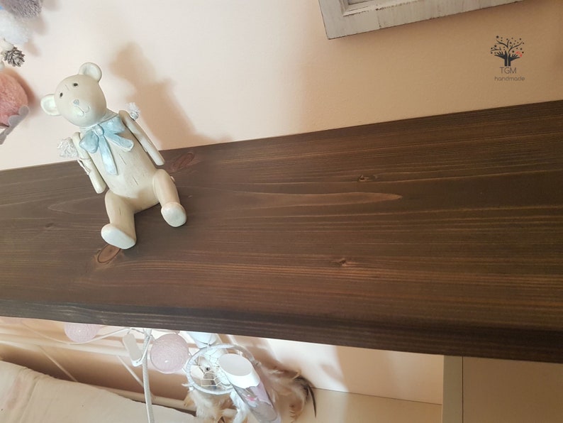 Puede incluir: Una figura decorativa de oso de peluche blanquecino con un lazo y detalles en azul claro. El oso est&aacute; colocado sobre una estanter&iacute;a de madera marr&oacute;n oscuro. La estanter&iacute;a est&aacute; montada en una pared rosa claro, creando una est&eacute;tica suave y acogedora.