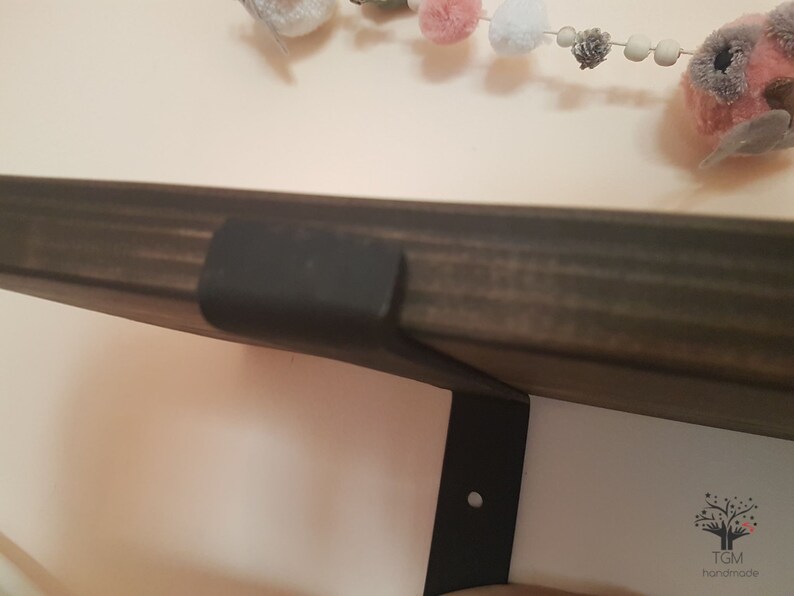 Puede incluir: Una estanter&iacute;a de madera te&ntilde;ida oscura con un soporte de metal negro fijado a una pared blanca. Una guirnalda decorativa con pompones rosas, grises y blancos y p&aacute;jaros de fieltro cuelga sobre la estanter&iacute;a. La estanter&iacute;a est&aacute; soportada por un soporte de metal negro.
