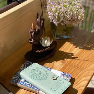 May include: Two small books with patterned covers, one green and one blue, are stacked on a wooden surface. The green book has the title "Traveller's Rest" on the cover. The blue book has the title "Paulo Coelho" on the cover. A small wooden incense holder with a hand-shaped design is in the background.