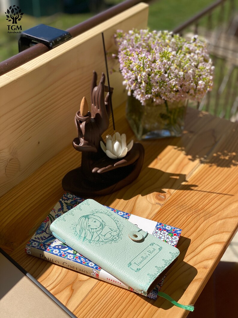 May include: Two small books with patterned covers, one green and one blue, are stacked on a wooden surface. The green book has the title "Traveller's Rest" on the cover. The blue book has the title "Paulo Coelho" on the cover. A small wooden incense holder with a hand-shaped design is in the background.