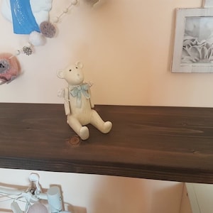 Puede incluir: Una peque&ntilde;a figura de oso de peluche blanquecino con una pajarita azul claro est&aacute; sentada en una estanter&iacute;a de madera marr&oacute;n oscuro. El oso est&aacute; sentado. Una guirnalda decorativa con pompones y una imagen enmarcada son visibles en el fondo.