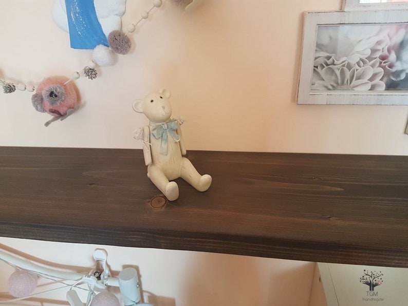 Puede incluir: Una peque&ntilde;a figura de oso de peluche blanquecino con una pajarita azul claro est&aacute; sentada en una estanter&iacute;a de madera marr&oacute;n oscuro. El oso est&aacute; sentado. Una guirnalda decorativa con pompones y una imagen enmarcada son visibles en el fondo.