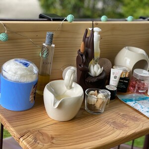 May include: A wooden shelf with a variety of spa and beauty products, including a bottle of ESPA body oil, a white ceramic bowl, a blue and white striped container, and a small glass jar filled with white beads. There are also several small bottles of essential oils and a white jar of cream.