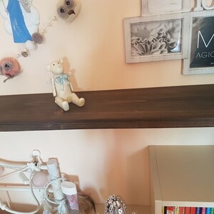 Puede incluir: Una peque&ntilde;a figura de oso de peluche blanquecino con un lazo azul claro est&aacute; sentada en una estanter&iacute;a marr&oacute;n oscuro. El oso tiene brazos y piernas peque&ntilde;os y est&aacute; colocado como si estuviera sentado. Hay objetos decorativos en el fondo.