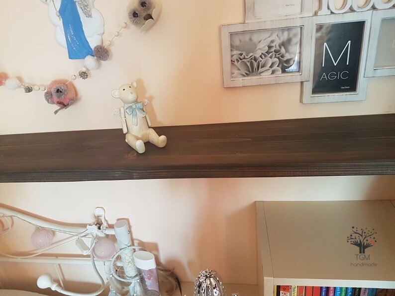Puede incluir: Una peque&ntilde;a figura de oso de peluche blanquecino con un lazo azul claro est&aacute; sentada en una estanter&iacute;a marr&oacute;n oscuro. El oso tiene brazos y piernas peque&ntilde;os y est&aacute; colocado como si estuviera sentado. Hay objetos decorativos en el fondo.