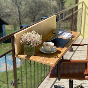 May include: A wooden desk with a laptop, a cup of coffee, and a vase of flowers. The desk is attached to a balcony railing. A brown leather office chair is in the foreground.