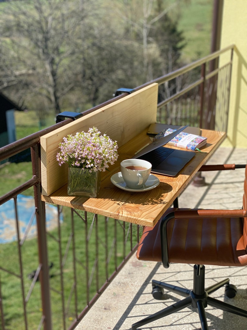 May include: A wooden desk with a laptop, a cup of coffee, and a vase of flowers. The desk is attached to a balcony railing. A brown leather office chair is in the foreground.
