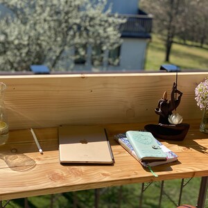 May include: A wooden desk with a laptop, two notebooks, a glass of water, a wooden incense holder, and a vase of pink flowers. The desk is on a balcony with a view of trees and a building in the background.
